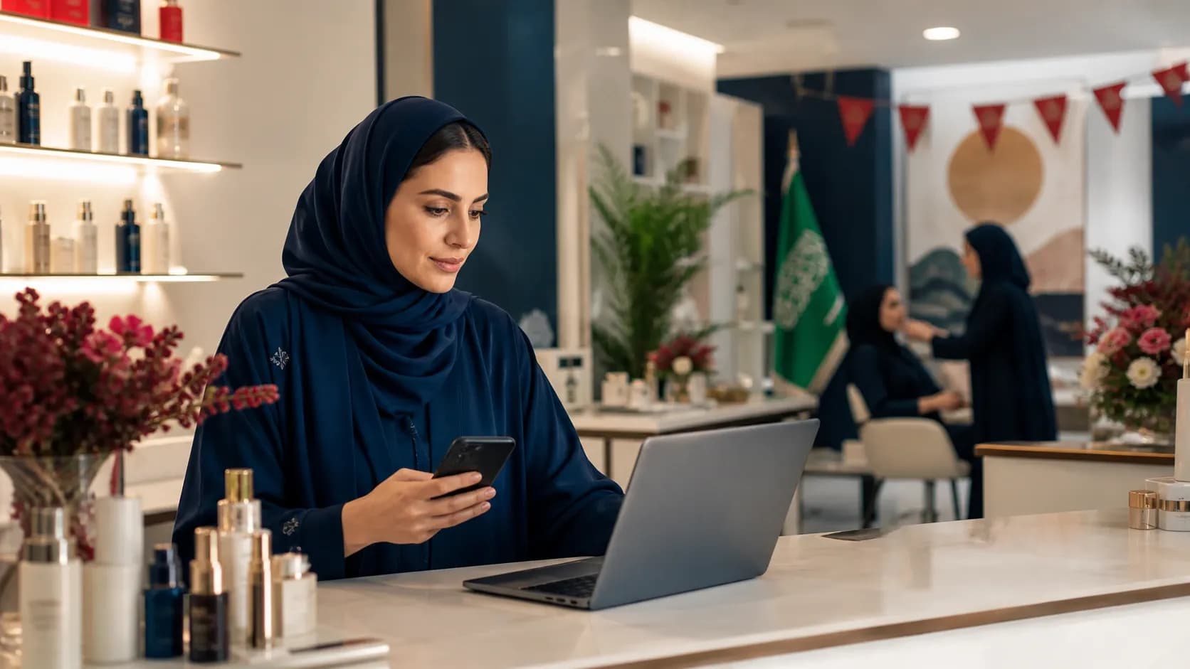 مديرة مركز عناية سعودي تراجع من مكتب الاستقبال مسارات حجز موسمية مختلفة على الهاتف والكمبيوتر المحمول داخل بيئة مرتبة